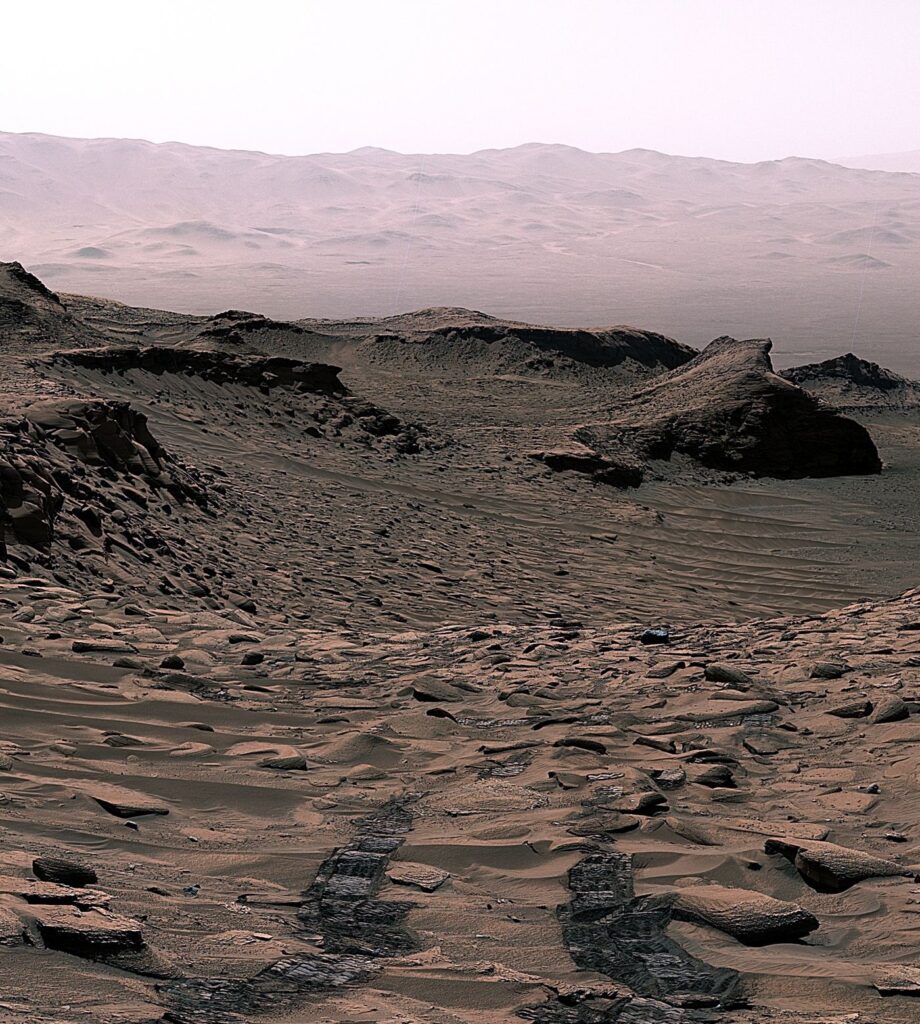 Flexible Mesh Wheels Could Help Mars Rovers Drive Farther A rover traversing the red rocky surface of Mars with layered terrain and a dusty horizon behind it.